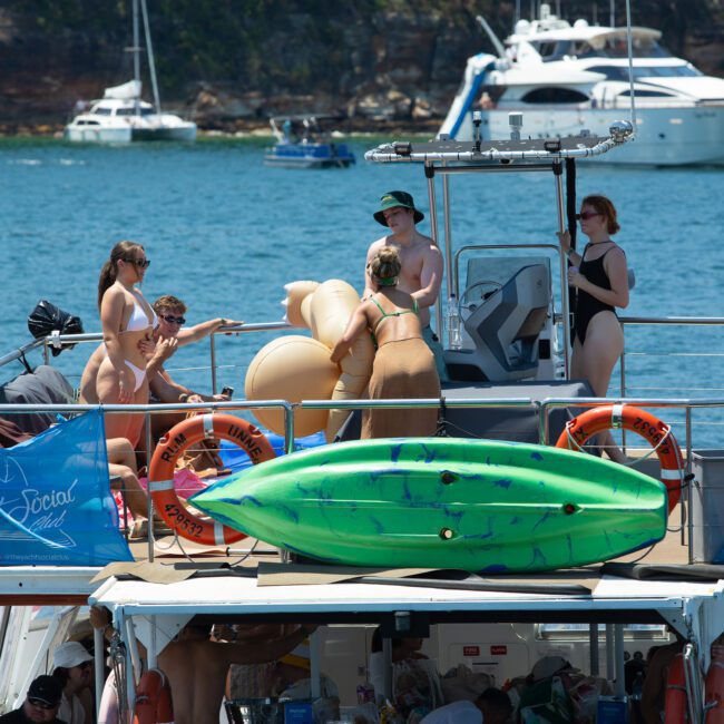 A group of people are gathered on a boat in a sunny, blue-watered location. Some are wearing swimsuits and interacting with a large, inflatable toy. A green kayak is secured to the roof of the lower deck, adding to the fun atmosphere. Other boats can be seen in the background.