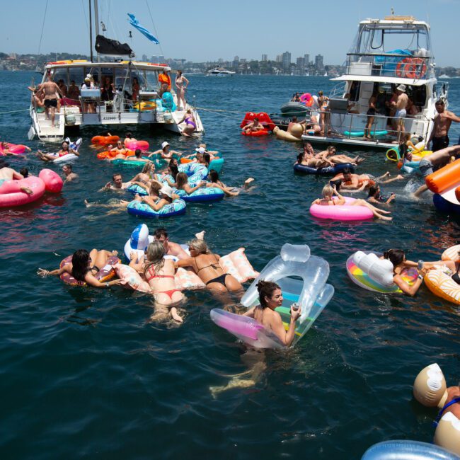 A lively scene of people lounging on colorful inflatable floats in a body of water, surrounded by boats. The group enjoys the sunny day, with various inflatables such as unicorns, donuts, and lounge chairs. The background showcases city buildings and a clear blue sky. Vibrant beach balls add to the fun atmosphere.