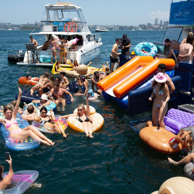 A lively scene of people in swimsuits enjoying a sunny day on the water. They are lounging on colorful inflatable floats and using a waterslide from a docked boat. A picturesque cityscape is visible in the background, with sailboats anchored nearby.