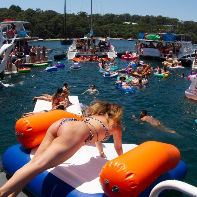 A lively scene with numerous people enjoying a sunny day on the water. Several boats are anchored closely, with people swimming, floating on colorful inflatables, and socializing. In the foreground, a woman in a bikini is climbing onto an inflatable unicorn.