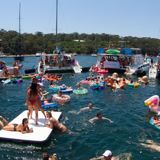 A lively scene of people enjoying a sunny day on a lake with boats and inflatables. Groups are swimming and relaxing on colorful flamingos and loungers, while others socialize on nearby paddleboards anchored close to each other.