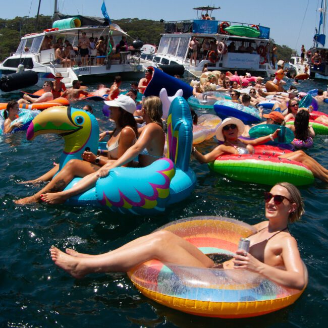 People enjoy a sunny day relaxing on various inflatable floats in the water, surrounded by boats. Some floats are colorful and shaped like animals or unicorns. Participants are chatting, sunbathing, and holding drinks, creating a lively and festive atmosphere.