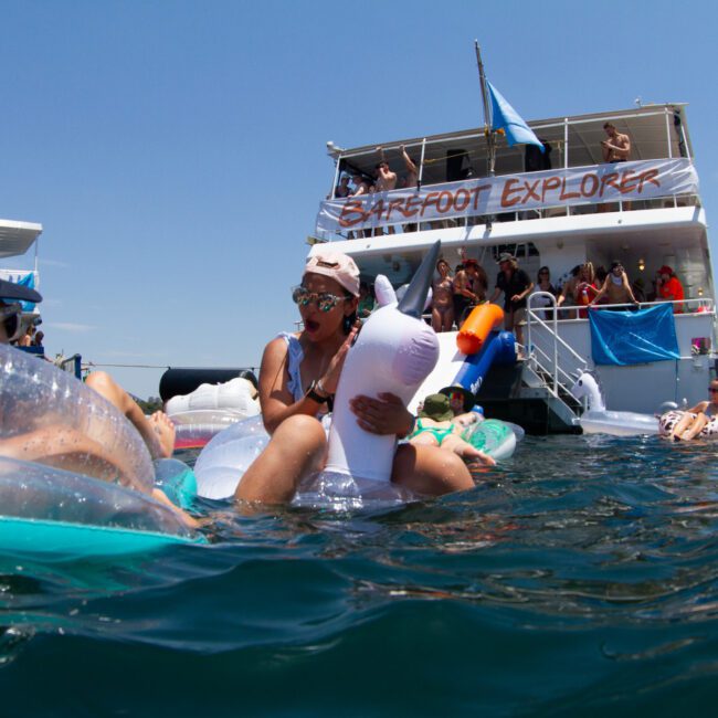 A lively scene of people enjoying a sunny day on the water. Many are floating on inflatable rafts and toys near a boat labeled "Barefoot Explorer." The background shows sailboats and participants. Everyone appears relaxed and having fun under the clear blue sky.
