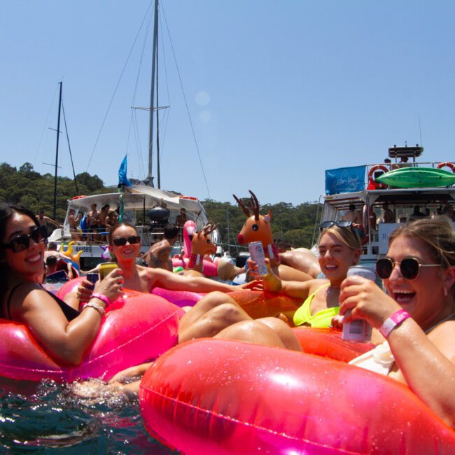 A group of people are relaxing on inflatable floaties in the water, holding drinks and smiling at the camera. Paddleboards and other people enjoying the sunshine can be seen in the background. The sun is shining, and it's a clear day with green trees visible in the distance.