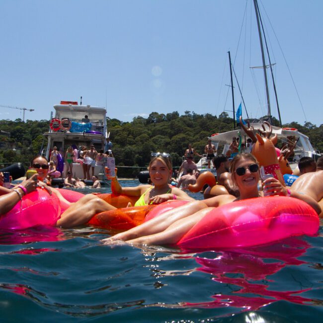 A group of people relax on inflatable floats in the water, holding drinks and smiling. In the background, boats are anchored, and more people can be seen enjoying the sunny day under a clear blue sky with trees lining the shore. The festive scene highlights a perfect summer getaway for friends and families alike.
