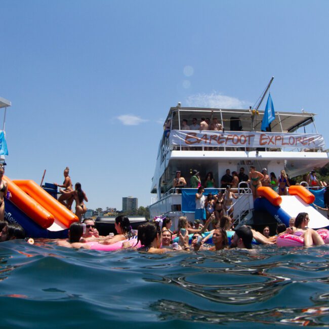 A lively outdoor scene showing a crowd of people swimming and enjoying the water near two boats. The boats are filled with additional people, slides, and inflatable toys. Tree-covered hills provide a picturesque backdrop under a clear sky, creating an idyllic summer setting.