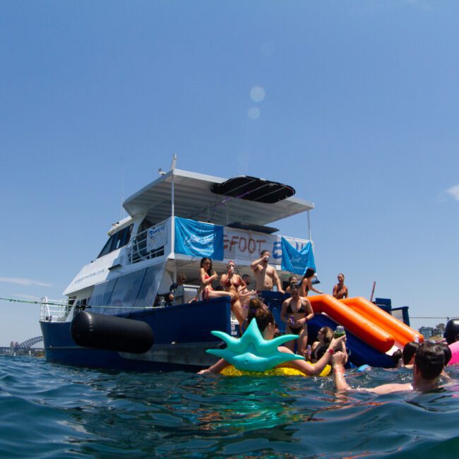 A group of people enjoying a sunny day on a blue boat with an orange slide, anchored near a city skyline. Some are swimming and using inflatable toys in the calm water, while others relax on the boat deck. The sky is clear with no clouds as they make lasting memories together.