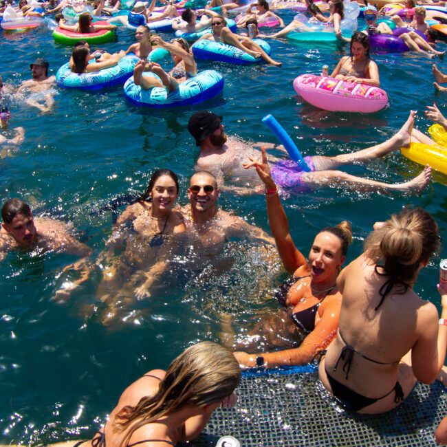 A lively group of people enjoy a sunny day on a lake, floating on colorful inflatables and swimming near boats. Everyone is in swimsuits, laughing, and having fun. Some hold drinks, while others lounge on the inflatables. Majestic mountains rise in the distance while dense green trees line the background.