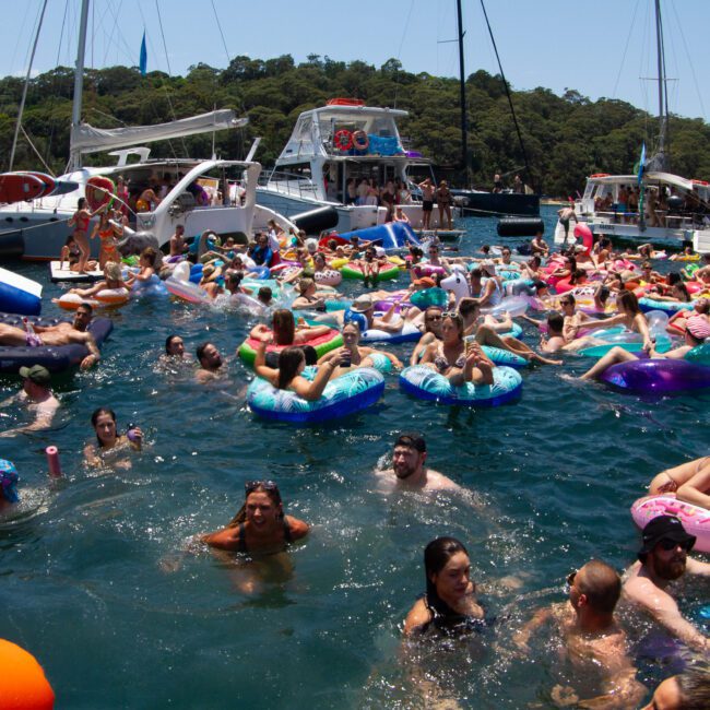 A large group of people enjoying a sunny day in the water, using various inflatable floats and lounging around. Boats are anchored in the background, creating a lively scene with people swimming, chatting, and relaxing. Trees surround the area while a clear sky stretches above.