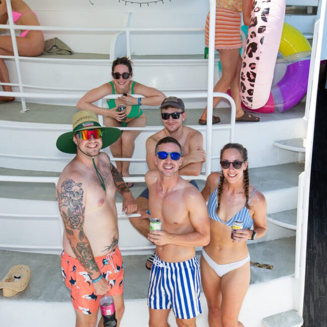 A group on a boat enjoys a sunny day. The foreground features three men and a woman in swimwear, holding drinks, while another man sits behind them. More people lounging in vibrant life vests are visible in the background along with inflatable floaties.