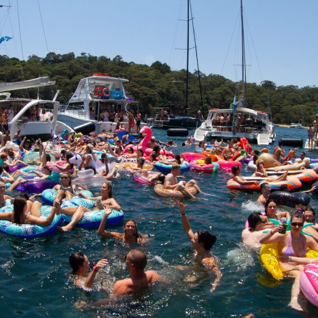 A large group of people are enjoying a sunny day on a lake, floating on inflatable rafts and toys. Colorful boats surround them, with green, tree-covered hills in the background. The festive scene includes lively music and laughter, making it a memorable day filled with fun.