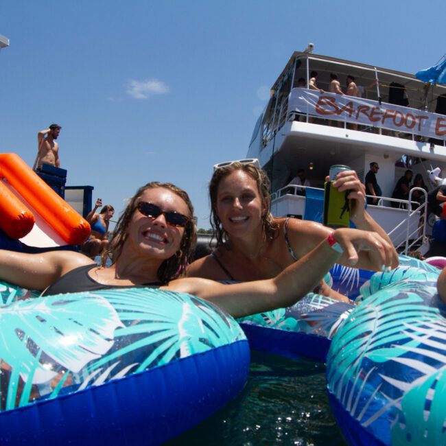 Two women are smiling and relaxing on inflatable floats adorned with tropical leaf patterns in the water. In the background, several people are on nearby boats, including one named "Barefoot Explorer." The sky is clear and the lively scene creates a sense of carefree adventure.