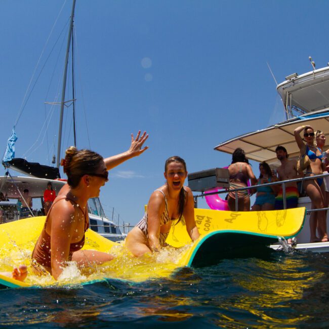 Two women laugh and play on a floating yellow mat in the water, while several people on boats in the background watch and take photos. The sky is clear and blue, creating a perfect backdrop for a sunny day at the marina.