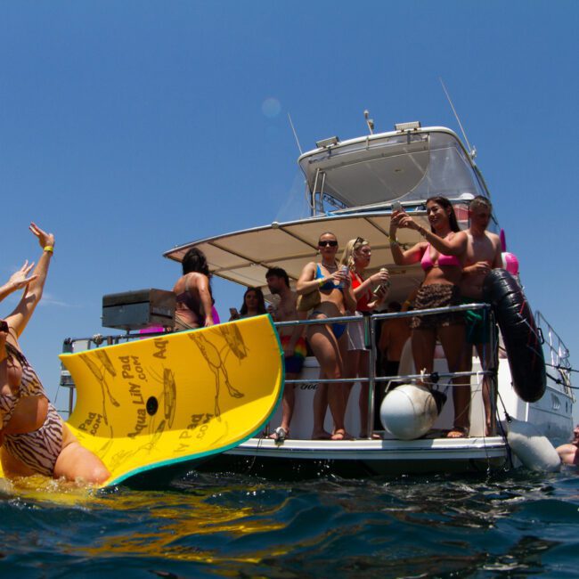 A group of people enjoying a sunny day on a boat and in the water. Two people lie on a floating mat, laughing and reaching towards individuals on the boat's deck. Others in swimsuits are taking photos, socializing, and admiring the serene sea.