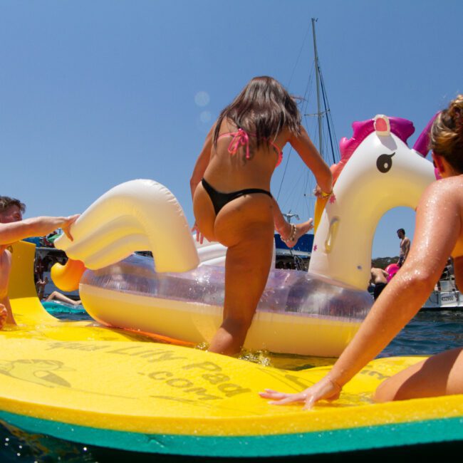 Three people enjoying a sunny day on the water. A woman climbs onto an inflatable unicorn, while another woman and a man relax on a floating mat nearby. Kayaks and clear blue skies are visible in the background.