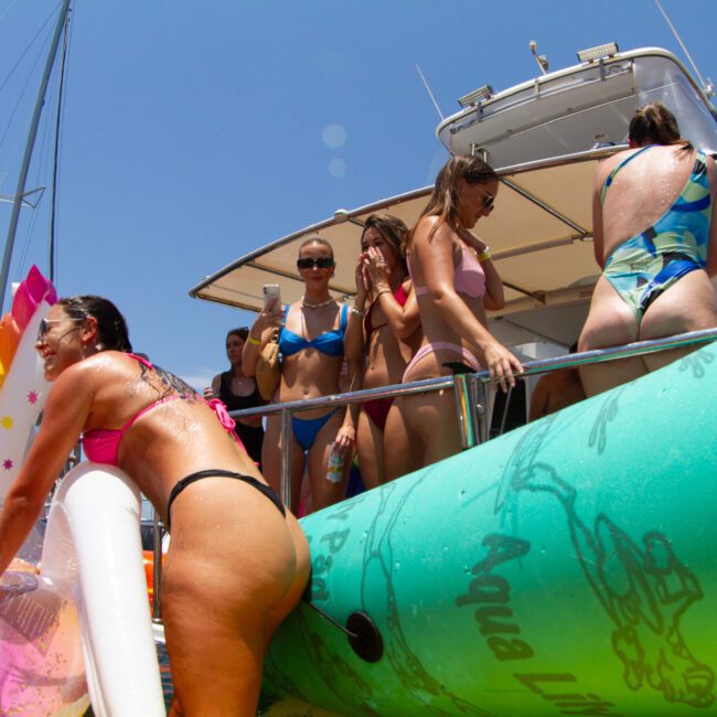 Several people in vibrant swimwear are enjoying a sunny day on a boat. Some are standing on the deck, while others float on a large inflatable tube in the water. They are engaged in lively conversation, holding drinks, and appear to be having a great time.