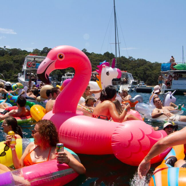A lively scene of people on a lake enjoying a sunny day on colorful inflatable pool floats, including a large pink flamingo and a unicorn. Several boats are anchored in the background, and everyone appears to be having fun, socializing while sipping beverages at sunset.