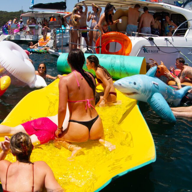 A lively scene of people enjoying a sunny day on the water. They are swimming, floating on playful inflatable toys (including a shark and a flamingo), and relaxing on a large yellow float near a yacht. Many others are socializing and mingling on the deck of the yacht.