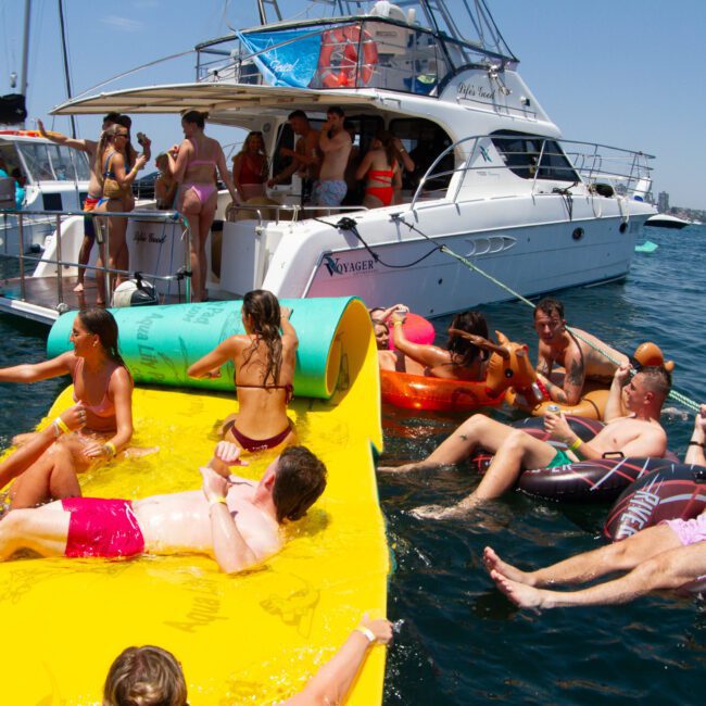 A group of people enjoying a sunny day on the water. Some are lounging on inflatable floats, while others relax on a large yellow mat floating near two boats. The mood is festive and relaxed, with everyone in swimsuits and engaging in conversation or soaking up the warm sunrays.