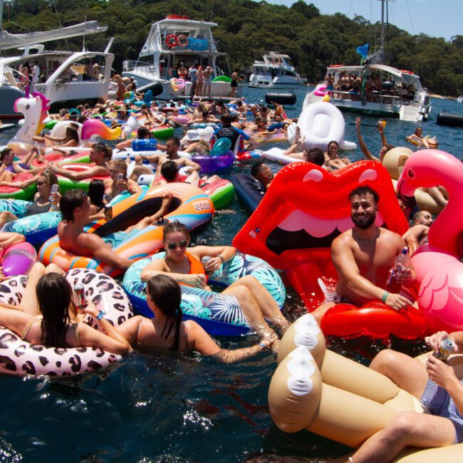 A large group of people is enjoying a sunny day on the water, floating on various inflatable devices like a flamingo, lips, and animals. Surrounded by boats in the background, they appear to be engaging in a lively, festive gathering with loads of excitement.