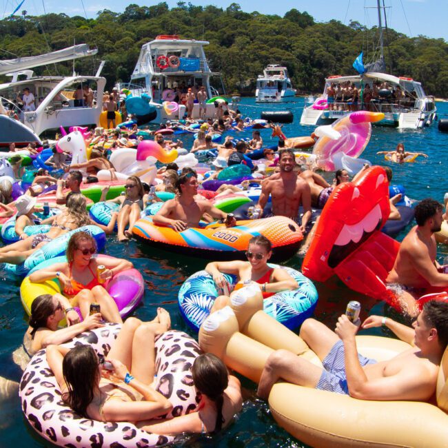A lively group of people enjoys a sunny day on the water, lounging on an assortment of colorful inflatable floats shaped like animals and food. Several boats are anchored nearby, creating a festive atmosphere with music, paddleboarding, drinks, and laughter.