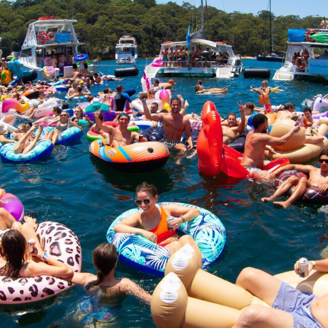 A lively scene of many people enjoying a sunny day on the water, floating on various colorful inflatable rafts, including shapes like flamingos and unicorns. Several yachts are anchored nearby, creating a festive, crowded atmosphere.