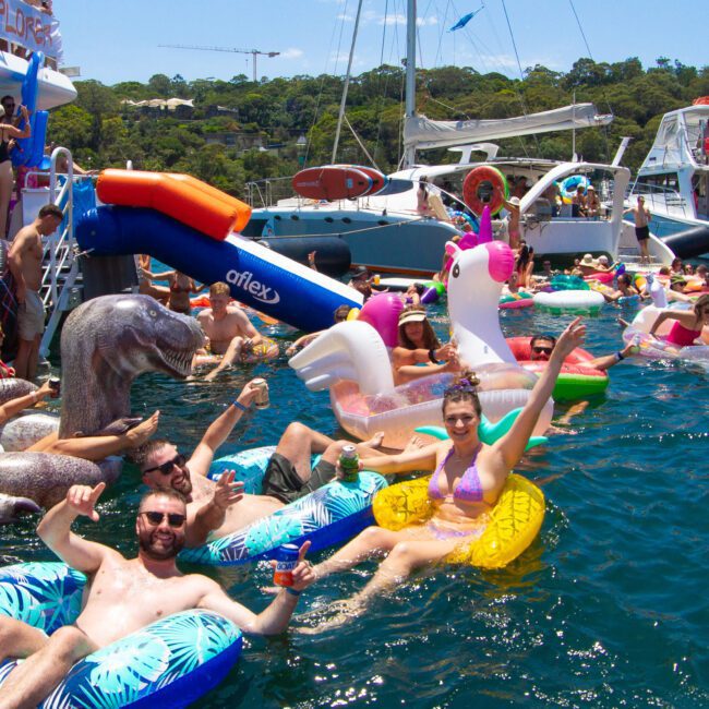 People enjoying a sunny day at a boat party, relaxing on various inflatable pool floats, including a swan and a unicorn. Others are gathered on boats docked nearby. The background shows lush greenery and clear blue skies, with music playing in the air.