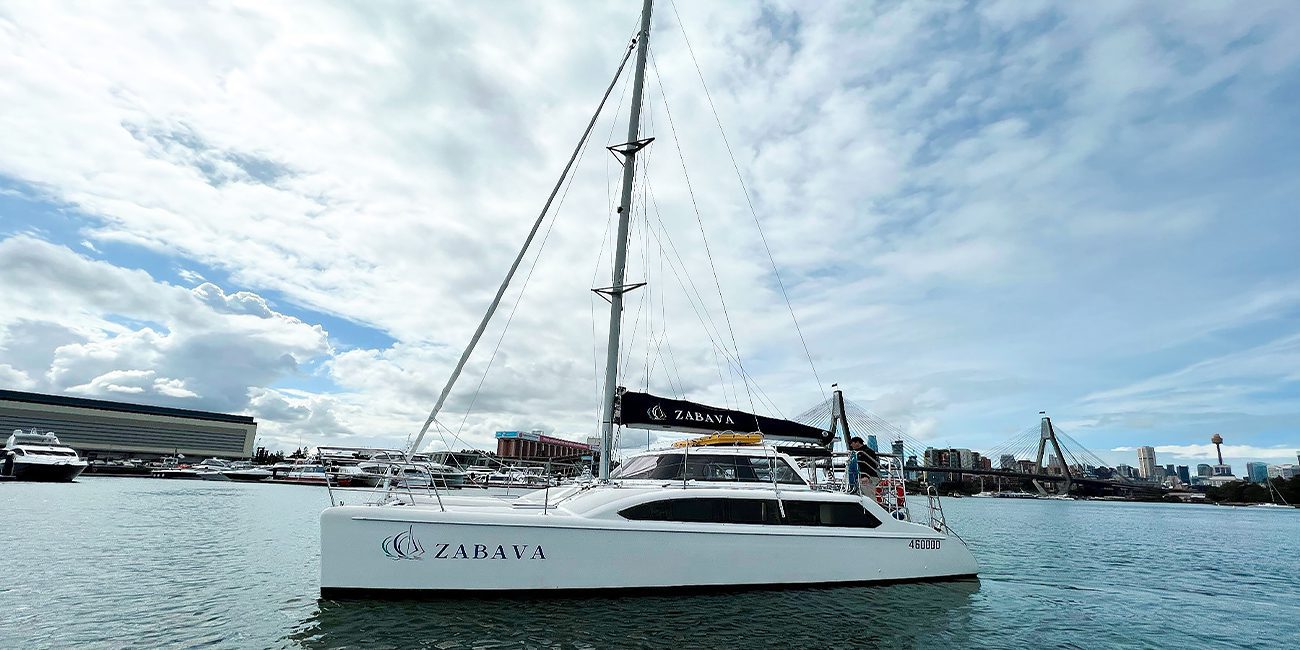 A white catamaran named "Zabava" is anchored in a calm harbor. The sky is partly cloudy, and several buildings and sailboats are visible in the background. The water reflects the sky and nearby structures, creating a tranquil scene.