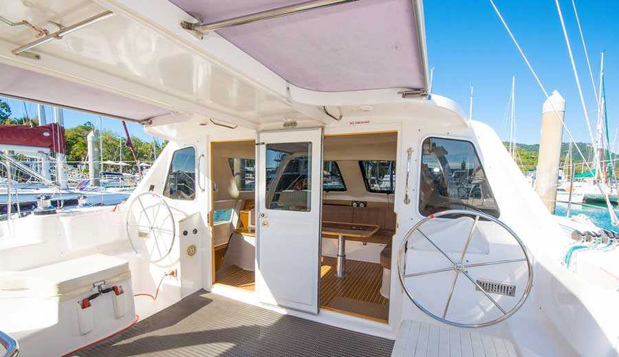 View of the cockpit and interior of a docked white catamaran. The cockpit, equipped with two steering wheels and a canopy for shade, offers a stunning marina view. Through open glass doors, the interior reveals a cozy seating area with a table. In the background, other boats are anchored under clear skies.