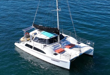 A white catamaran named "CABARET" floats on clear blue water. The boat features several solar panels on its roof, a black sail cover, and red storage units on the bow. With a sleek and modern design, it looks ready for an adventure.