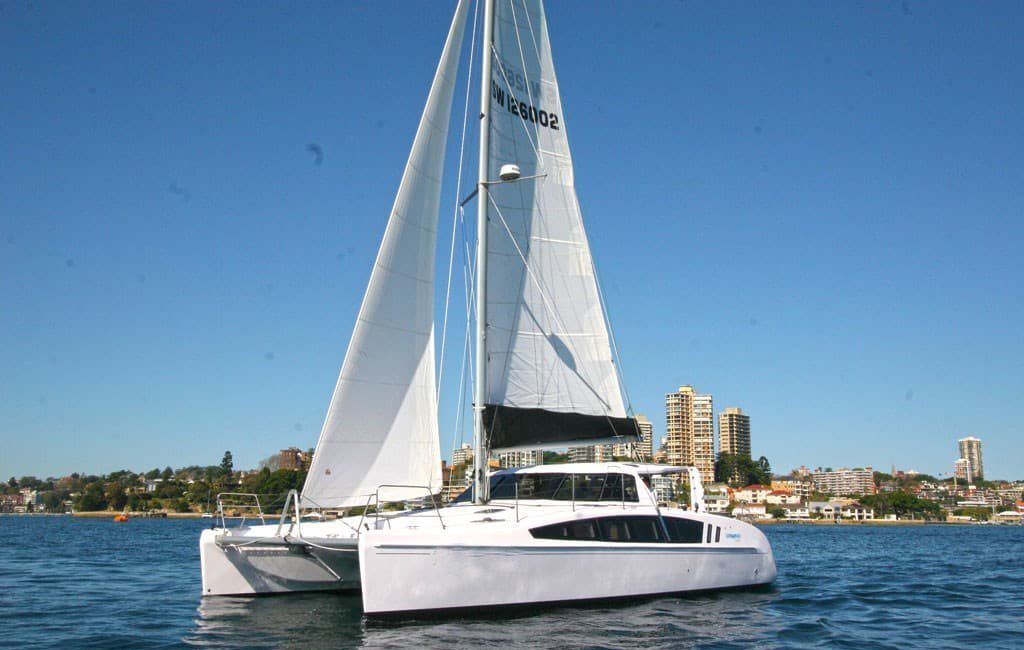 A white sailboat with tall sails glides on a calm body of water. In the background, there are picturesque buildings and residences visible along the shoreline under a clear blue sky.