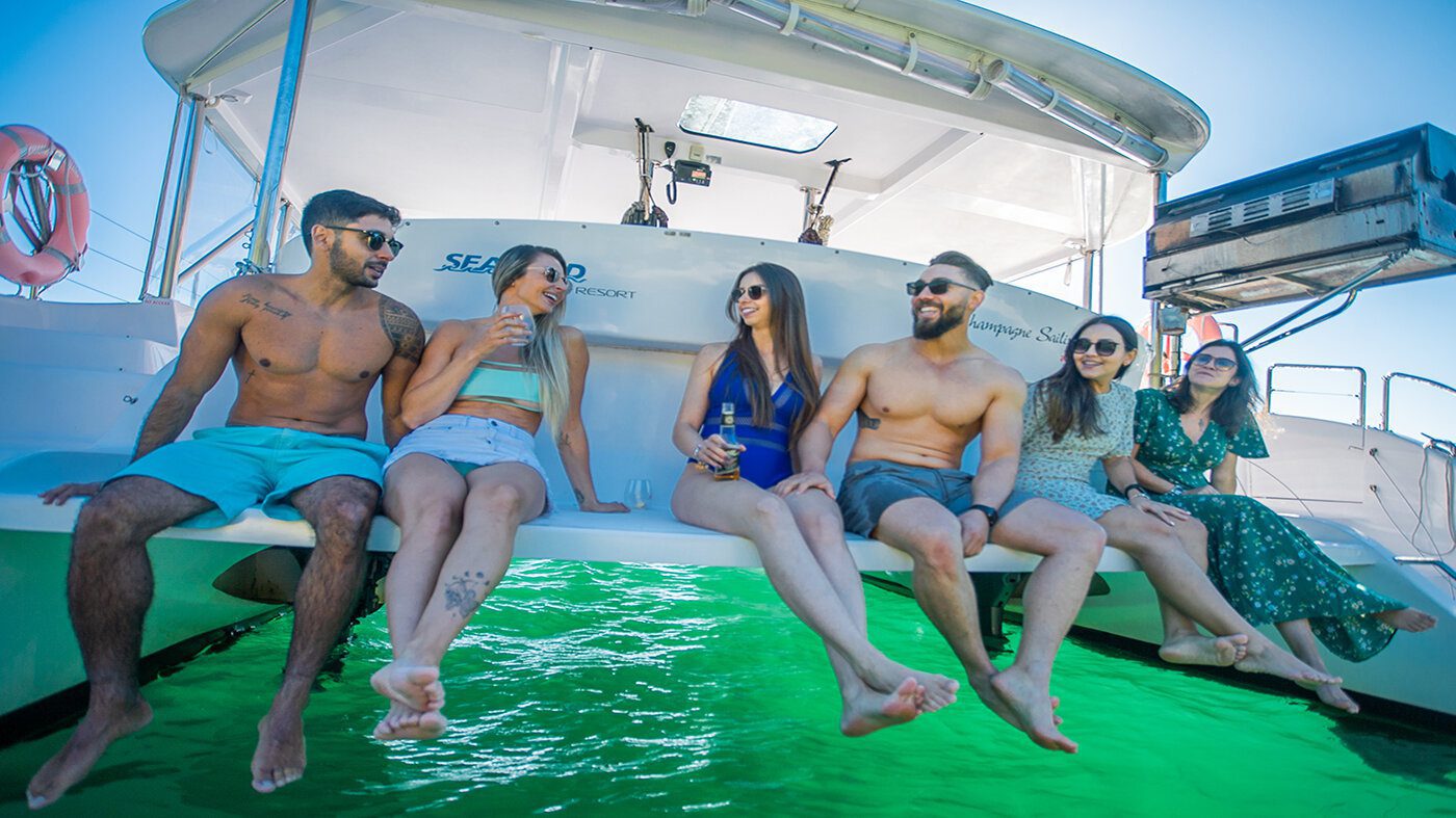 A group of six people, three men and three women, are sitting at the edge of a boat with their feet dangling over the bright green water. They are in swimsuits and casual summer attire, smiling, chatting, and holding drinks under a clear blue sky.