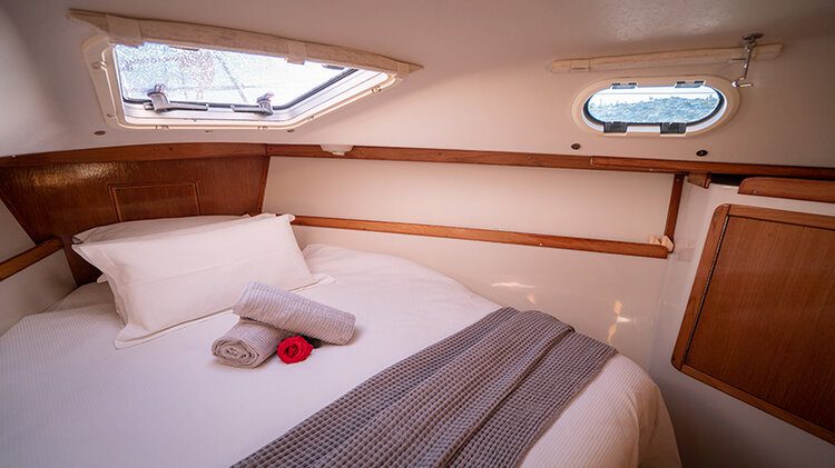 A cozy cabin bedroom featuring a neatly made bed with white linens, a gray throw blanket, and two rolled towels with a red rose placed on top. The room has two small windows allowing natural light to enter, wooden accents, and a compact storage shelf perfect for keeping your things organized.