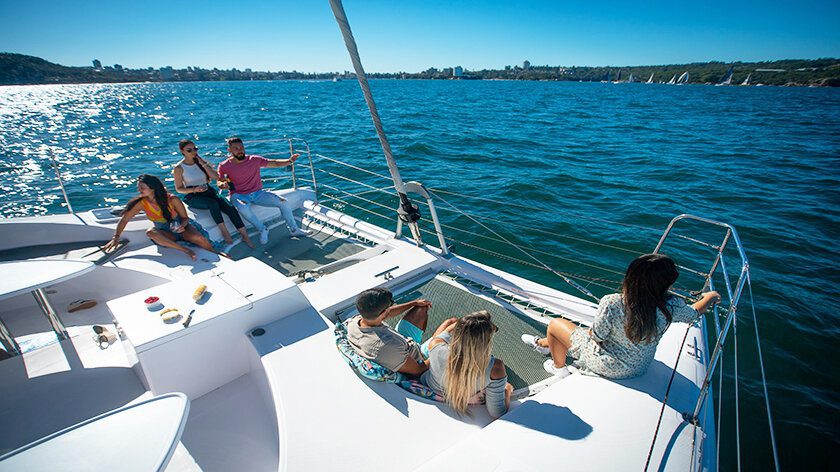 Five people are relaxing and socializing on a large white sailboat on a sunny day. The sailboat glides gently across a calm, expansive body of water with a distant shoreline and scattered boats in the background.