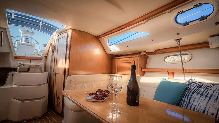 The cozy cabin interior of a sailboat features a wooden table set with two wine glasses, a bottle of wine, and a plate of grapes. Cream-colored cushioned seating enhances the space, complemented by portholes and a hatch letting in natural light. Adjacent steps lead to the teak deck outside.
