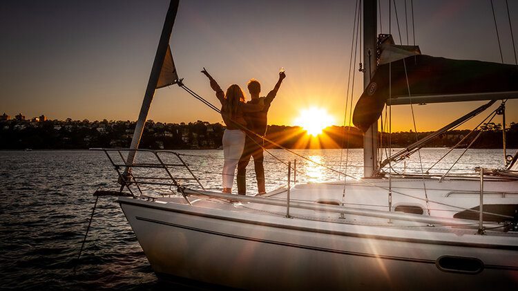 A couple stands on the deck of a sailboat during sunset, with arms raised and a glass in hand. They are facing towards a picturesque shoreline as the sun dips below the horizon, casting a warm golden glow over the serene water.