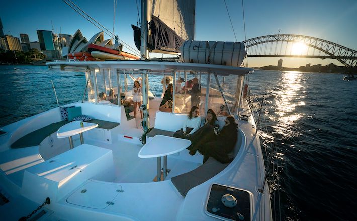 A group of people relax and enjoy the sunset on a sailing catamaran in a harbor. The Sydney Harbour Bridge and city skyline are visible in the background. The deck has seating areas, and life vests are stored overhead. A gentle breeze adds to the serene atmosphere as the setting sun casts a warm glow over the scene.