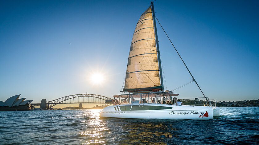 A white sailboat labeled "Champagne Sailing" glides on the water with a group of people aboard. In the background, the iconic Sydney Harbour Bridge and Sydney Opera House are visible under a clear blue sky as the sun sets, enhancing the serene ambiance.