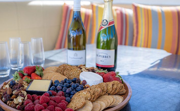 A charcuterie board with a variety of crackers, cheese, grapes, strawberries, and mixed nuts is placed on a table. Behind the board are two bottles of wine and a few empty wine glasses. The background features colorful, striped cushions on a cozy seating area.