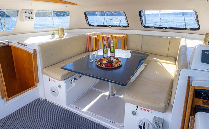 Interior of a boat with a seating area featuring beige cushioned benches, a rectangular table with a platter of snacks, four colorful striped pillows, and windows revealing a scenic water view. The table is decorated with glasses, a bottle of sparkling drink, and cutlery. Subtle marine-themed decor enhances the ambiance.