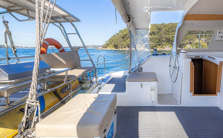 A spacious deck of a boat on a sunny day, featuring cushioned seating, a stainless steel railing, life jackets, and a life ring. The open water and shoreline with trees are visible in the background. Various navigational equipment and a steering wheel are also present.