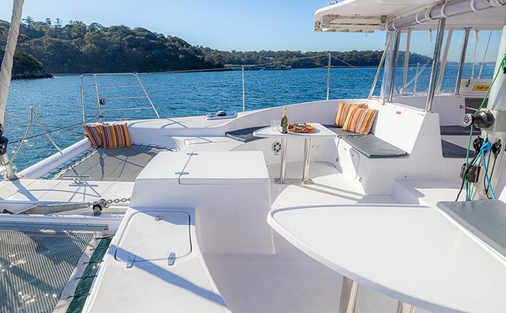 A spacious deck of a white catamaran features cushioned seating around a small table with fruit, an open area with netting, and scenic ocean views in the background. Trees and shoreline are visible in the distance under a clear blue sky, perfect for sunbathing.