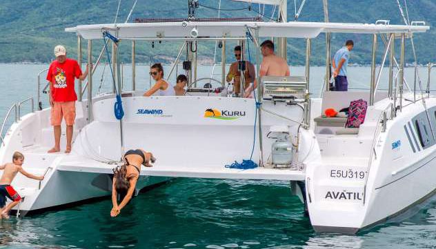 People are savoring a sunny day on a white catamaran named "AVATIU." Some individuals are sitting and standing on the deck, while a woman is diving into the refreshing water and a child stands at the side of the boat, extending a hand to another person.