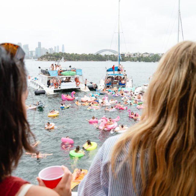 People on a boat enjoying a lively scene with numerous individuals floating on inflatable rafts in the water. The background features additional boats and a distant city skyline under a cloudy sky. Experience this vibrant atmosphere with Luxury Yacht Rentals Sydney from The Yacht Social Club.