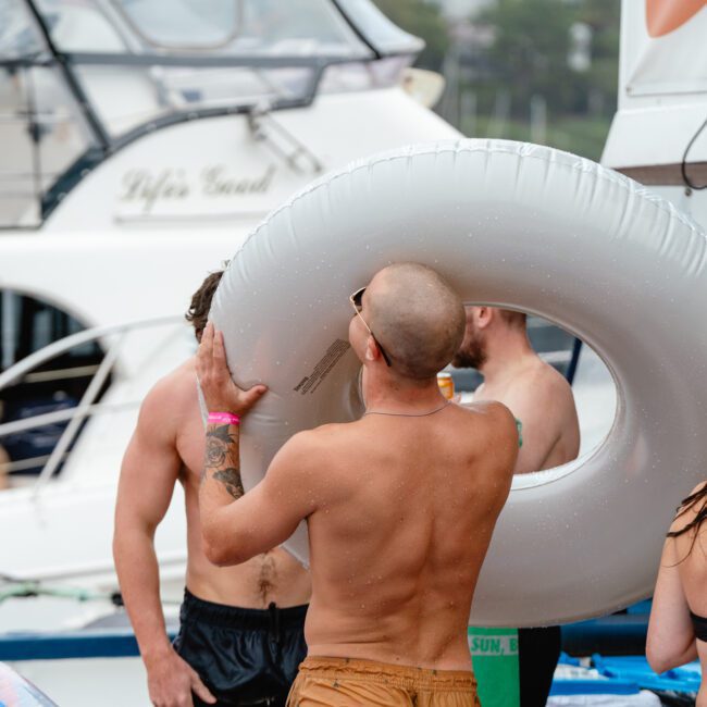A group of people enjoying a boat party with The Yacht Social Club Sydney Boat Hire. A shirtless man holds a large white inner tube over his head, while others in swimsuits are visible. Boats are in the background, and a banner appears on the right side.