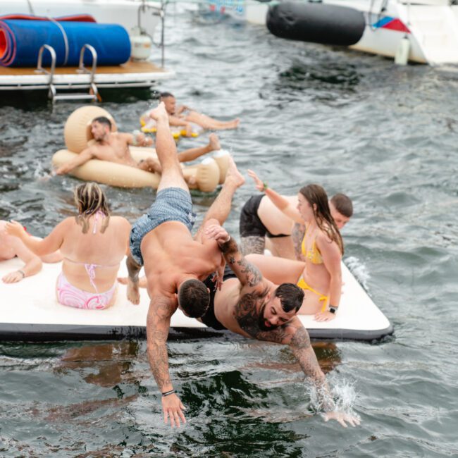 A group of people enjoys a day on the water with some swimming and lounging on floats. Two people are playfully wrestling on a floating mat while others relax and chat. Boats from The Yacht Social Club are docked nearby, creating a lively and fun atmosphere.