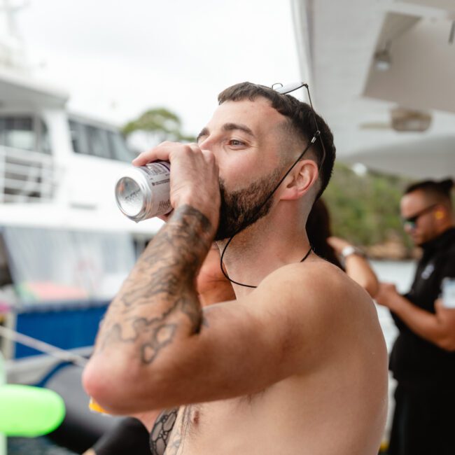 A shirtless man with tattoos is sipping from a can on a boat. He is wearing sunglasses on his head. Behind him, a person in a black shirt and another boat can be seen. The setting appears to be a lively outdoor gathering, typical of Boat Parties Sydney by The Yacht Social Club.