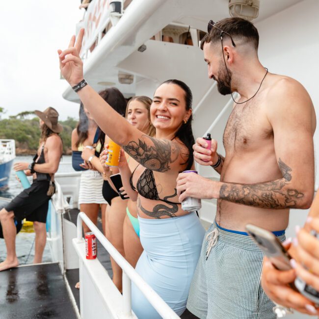 A group of people stand on a boat deck, enjoying a lively atmosphere. A woman in the center in a black swim top and light blue leggings smiles and throws a peace sign. A man next to her in swim trunks holds a beverage. Other passengers are in the background, all part of The Yacht Social Club Sydney Boat Hire event.