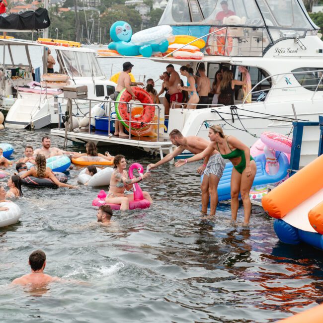 A group of people are enjoying a boat party hosted by The Yacht Social Club Sydney Boat Hire. Several are in the water using inflatable toys, while others are on boats. Two people on a large float are helping someone out of the water. The background shows various boats and a lush, green shoreline.