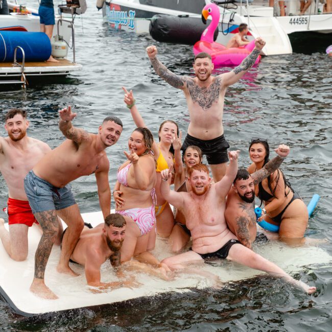 A group of people, some with tattoos, in swimwear enjoy themselves on a float in the water. They are smiling, raising their arms, and posing for the photo. In the background, there are boats from The Yacht Social Club Sydney Boat Hire and a large pink flamingo float.